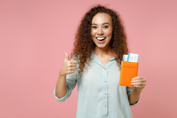 Traveler tourist black african woman in blue shirt hold passport tickets show thumb up gesture isolated on pastel pink background. Passenger travel abroad weekends getaway. Air flight journey concept