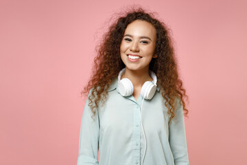 Young black african american fun student cute happy smiling positive friendly curly woman 20s in blue casual shirt wearing headphones looking camera isolated on pastel pink background studio portrait