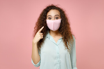 Young black african curly woman in blue shirt point index finger on face mask to safe from coronavirus virus covid-19 during pandemic quarantine isolated on pastel pink background studio portrait