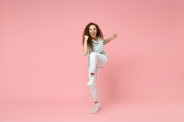 Fototapeta na wymiar Full length young black african happy overjoyed excited friendly curly woman in blue shirt stand do winner clench fist with raised up leg celebrate isolated on pastel pink background studio portrait.