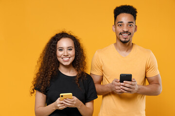 Young couple two friends together family african man woman 20s in black t-shirt holding using mobile cell phone chatting in social network dating isolated on yellow color background studio portrait.