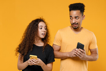Young couple two friends together family african curious jelaous woman man in black t-shirt hold using mobile cell phone peeping boyfriend message isolated on yellow color background studio portrait.