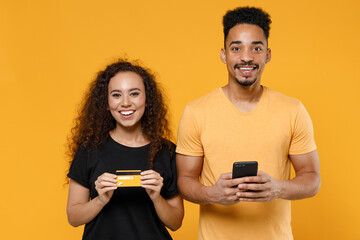 Young couple two friends together family african smiling happy man woman in black tshirt hold mobile cell phone credit bank card shop online look camera isolated on yellow background studio portrait