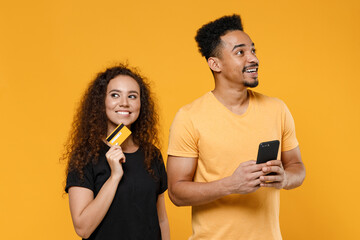 Young couple two friends together family african dreamful man woman 20s in black t-shirt holding mobile phone credit bank card shop online look aside isolated on yellow background studio portrait.