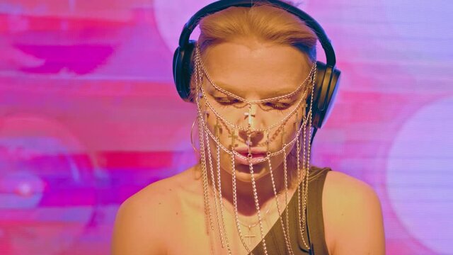 A cool and stylish female DJ of European appearance with blonde hair and a fashionable face mask made of Christian crosses works in a nightclub behind the turntables and creates dance music.