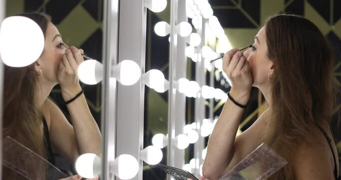 Beautiful young woman doing makeup in the dressing room in front of the mirror. Model uses a brush to apply shadows on the eyelids of the eyes
