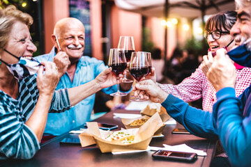 Senior people toasting wine at restaurant bar wearing open face masks - New normal life style...