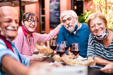 Happy senior friends taking selfie at restaurant with open face mask - Retired people having fun together at winebar after lockdown reopening - Positive elderly life style concept on vivid warm filter