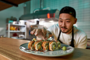Professional sushi chef wearing protective gloves decorating rolls served on plate at commercial kitchen
