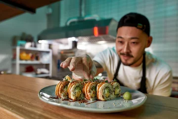 Fotobehang Professional sushi chef wearing protective gloves decorating rolls served on plate at commercial kitchen © Svitlana