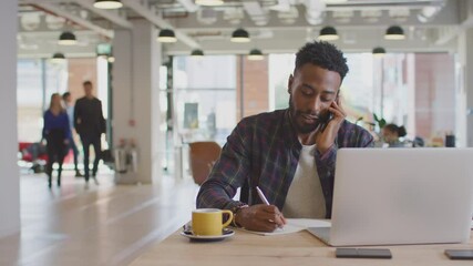 Dolly shot of casually dressed young businessman laughing on phone call sitting at desk in modern open plan start up office with colleagues in background - shot in slow motion - Powered by Adobe