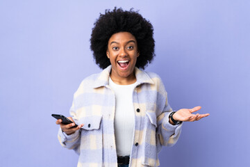 Young African American woman using mobile phone isolated on purple background with shocked facial expression