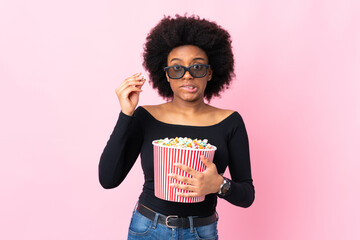 Young African American woman isolated on pink background with 3d glasses and holding a big bucket of popcorns