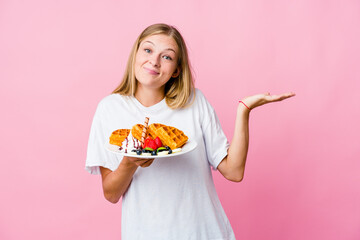Young russian woman eating a waffle isolated doubting and shrugging shoulders in questioning...