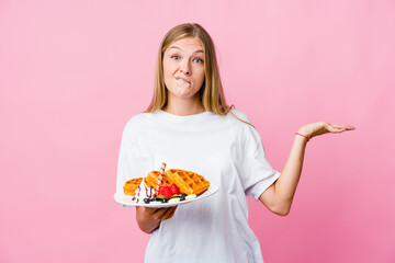 Young russian woman eating a waffle isolated confused and doubtful shrugging shoulders to hold a copy space.