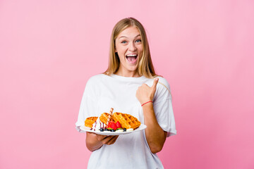 Young russian woman eating a waffle isolated surprised pointing with finger, smiling broadly.