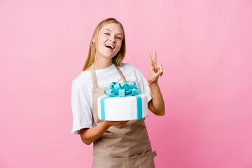 Young russian baker woman holding a delicious cake showing victory sign and smiling broadly.