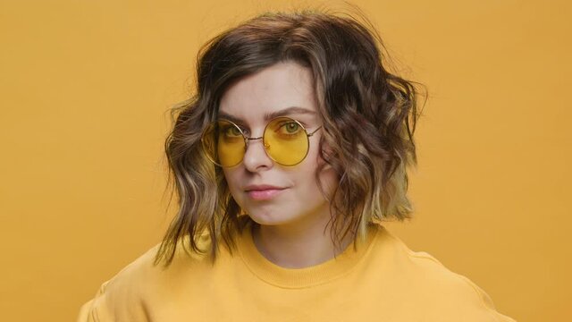 Happy Young Woman Smiles and Turns her head towards look on camera in Sunglasses on Yellow background slow motion. Glamorous blonde girl with curly hair. Positive emotions. Fashion. Freedom. Lifestyle