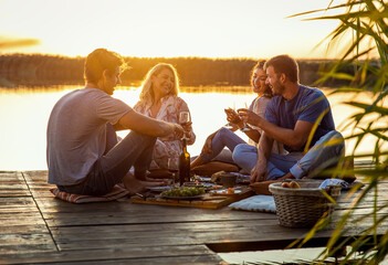 Group of friends having fun on picnic near a lake, sitting on pier eating and drinking wine.