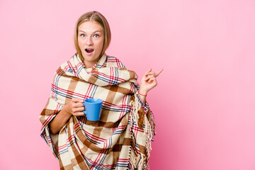 Young russian woman wrapped in a blanket drinking coffee pointing to the side