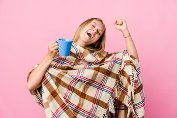 Young russian woman wrapped in a blanket drinking coffee celebrating a special day, jumps and raise arms with energy.