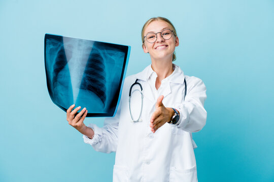 Young russian doctor woman holding a bone scan on blue stretching hand at camera in greeting gesture.