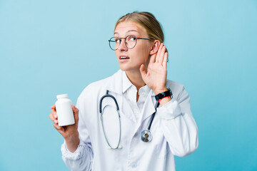 Young russian doctor woman holding pills bottle on blue trying to listening a gossip.
