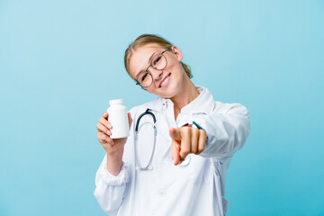 Young russian doctor woman holding pills bottle on blue cheerful smiles pointing to front.