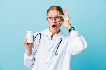 Young russian doctor woman holding pills bottle on blue keeping eyes opened to find a success opportunity.