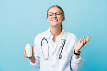 Young russian doctor woman holding pills bottle on blue joyful laughing a lot. Happiness concept.