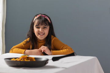 A SMILING YOUNG GIRL SITTING IN FRONT OF CAMERA WITH FOOD KEPT ON TABLE	