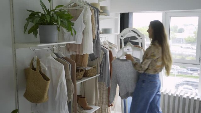 Side view fashionable young woman getting dressed in wardrobe near panoramic window and pot plants on shelf. Lade stylist chose and trying clothes from rack in open cupboard at home