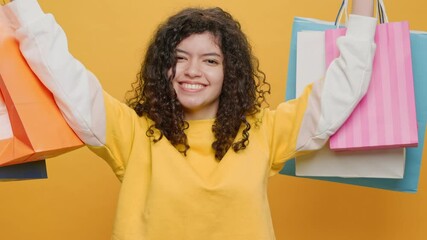 Happy Young Brazilian Woman from Shopping Bags Dancing in front Rhythmically Moves her Arms Up Down, Looks at Camera Fashion. Sales in Malls. Black Friday. Portrait Glamorous Girl on Yellow Background