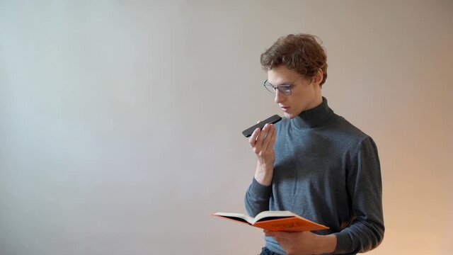 A young guy with glasses reads a book and listens to the sound on his smartphone