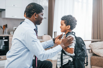 Obraz premium African american father with his son is indoors at home. Man helps child to wear backpack