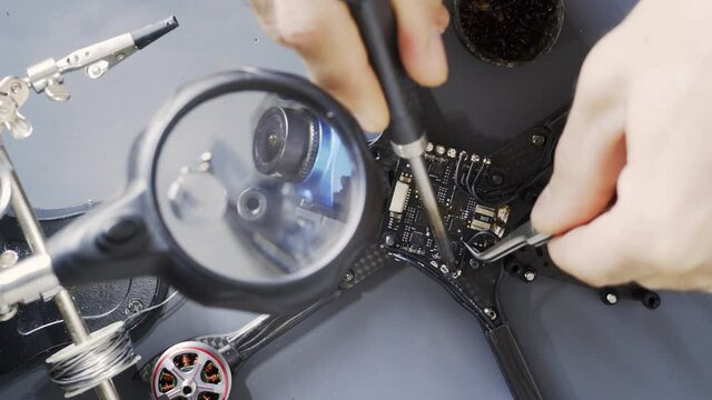 Detail shot skilled technician hands solder wire to chip of reassembled modern drone on grey plastic tray close view through magnifier glass. Concept DIY electronic project