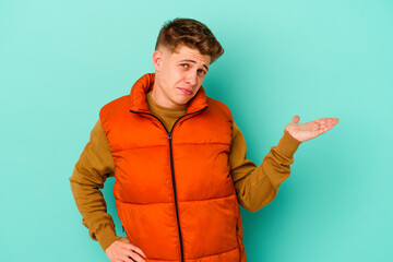 Young caucasian man isolated on blue background doubting and shrugging shoulders in questioning gesture.