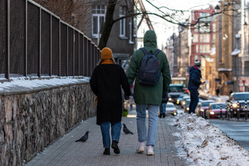a couple of people holding hands walking along the sidewalk along the side of the street, view from behind