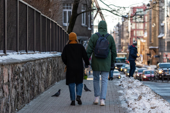 A Couple Of People Holding Hands Walking Along The Sidewalk Along The Side Of The Street, View From Behind