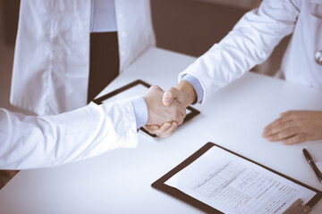 Unknown doctors are shaking their hands finishing discussion about patient's diagnosis, sitting at the desk and using a clipboard, close-up. Medical help, insurance in health care, medicine concept