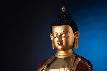 The top part of a gilded statue of Medicine Buddha, in traditional clothes with ushnisha on the head.  Isolated on black background with blue light spot.