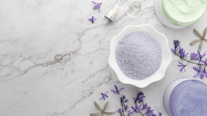 Obraz na płótnie Canvas Natural spa body scrub products set. Sea salt in bowl with sugar scrub, moisture cream, essential oils and violet flowers on marble table. DIY skin care routines for healthy skin. Flat layout.
