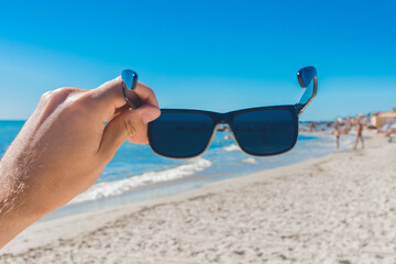 A young man's hand holds sunglasses against a sea beach