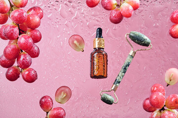 Organic cosmetics with grape seed oil and jade massage roller. Zero Waste Winemaking. Skincare product bottle, grapes and water drops on pink background