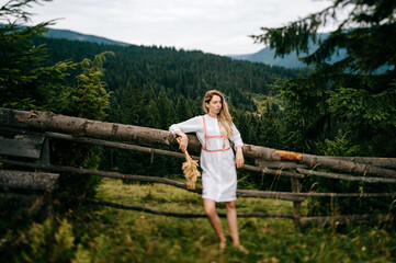 Fototapeta na wymiar Young attractive blonde girl in white dress with embroidery posing with spikelets bouquet near wooden fence