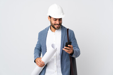 Young architect man with helmet and holding blueprints isolated on white background sending a message with the mobile