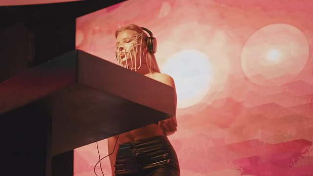 A cool and stylish female DJ of European appearance with blonde hair and a fashionable face mask made of Christian crosses works in a nightclub behind the turntables and creates dance music.