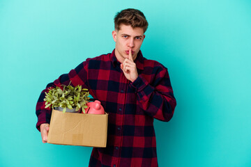 Young caucasian man moving holding a box isolated on blue background keeping a secret or asking for silence.