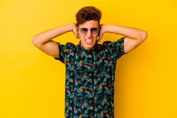 Young caucasian man wearing a Hawaiian shirt isolated on yellow background covering ears with hands trying not to hear too loud sound.