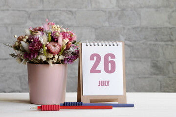 july 26. 26th day of the month, calendar date.A delicate bouquet of flowers in a pink vase, two pencils and a calendar with a date for the day on a wooden surface